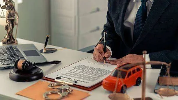 Advogado assinando contrato em mesa com laptop, malhete, algemas, balança, estátua da justiça e carro
