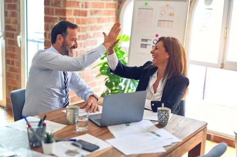 Dois profissionais de negócios sorrindo e dando um 'high-five' em um escritório, com laptop e documentos na mesa